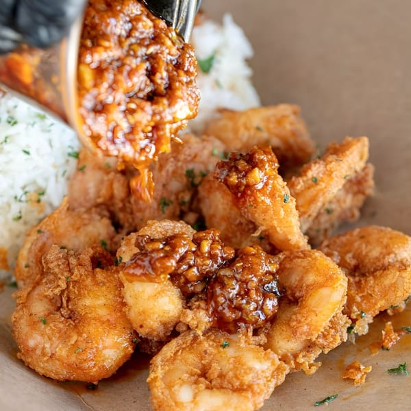 Golden fried shrimp being drizzled with a rich, dark sauce, served with white rice.